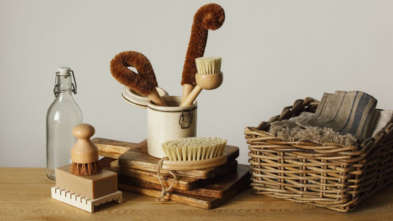 A set of sustainable coconut bristle cleaning tools on a wooden table. A woven wicker basket of rustic cleaning clothes beside it. 