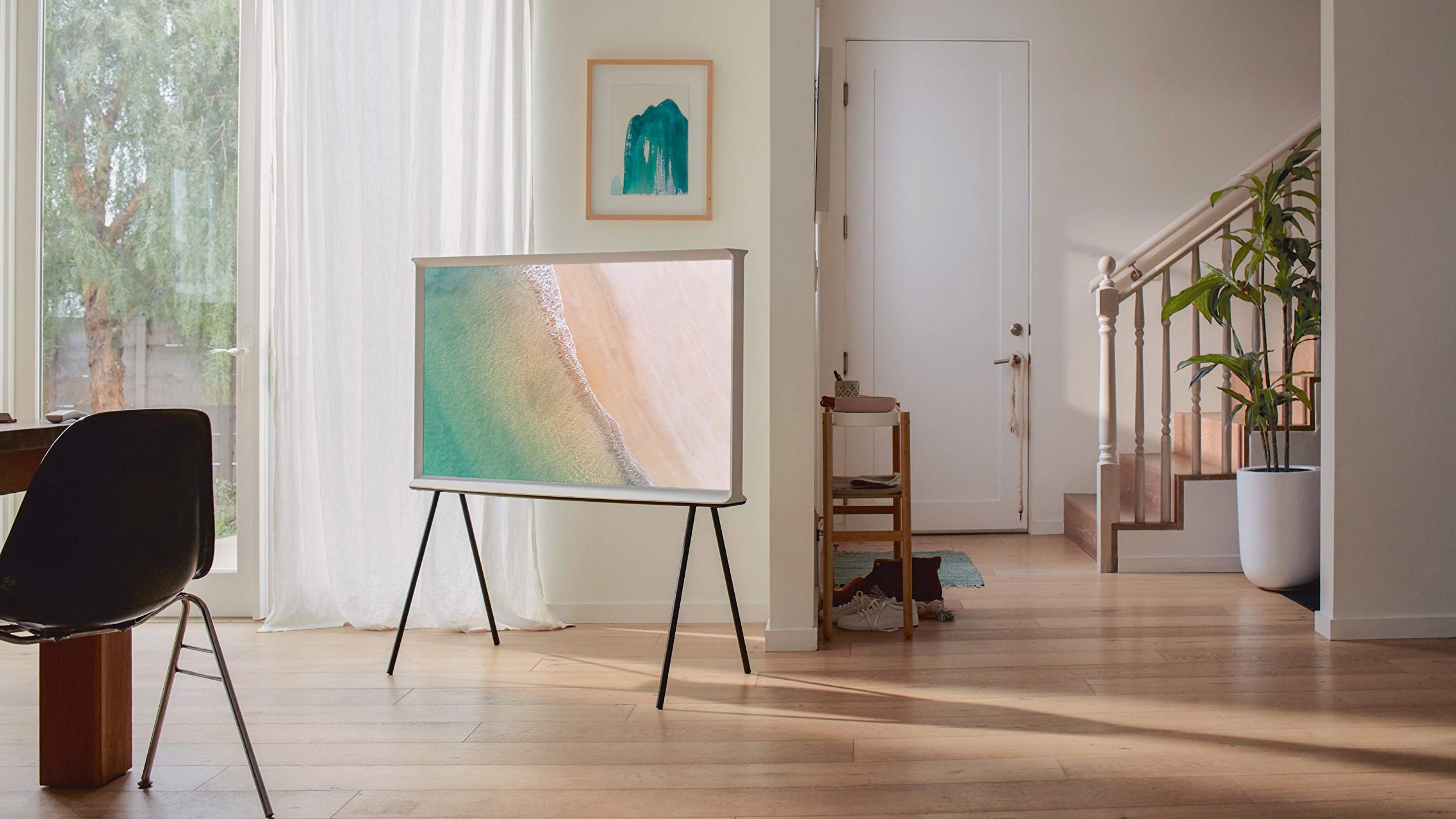 A naturally lit living room with a flat-screen, canvas-looking TV standing on two wrought iron inverted-V legs on wooden flooring, decorated with minimal furniture and plants.