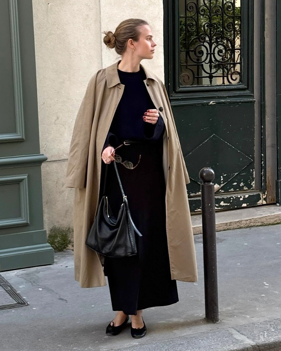 Danish fashion creative Clara Dyrhauge poses in Paris in front of a green door wearing a trench coat, black maxi dress, and black ballet flats