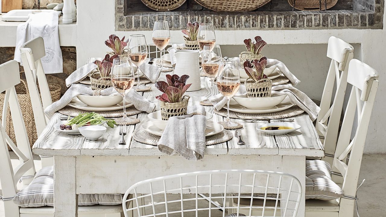 Dining table set with white tableware, rattan round placemats, houseplants, and drink poured in wine glass