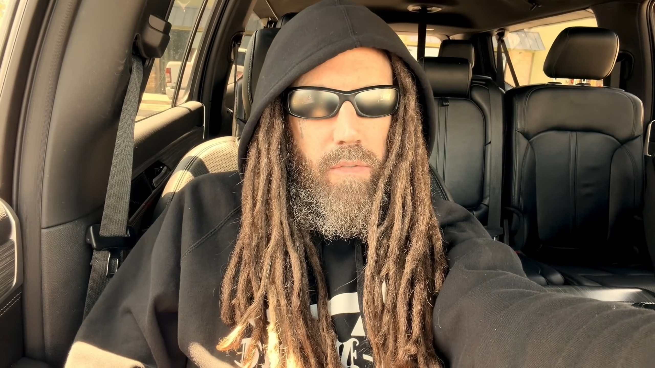 A person with long dreadlocks and a beard sits in a car wearing a black hoodie and sunglasses. T