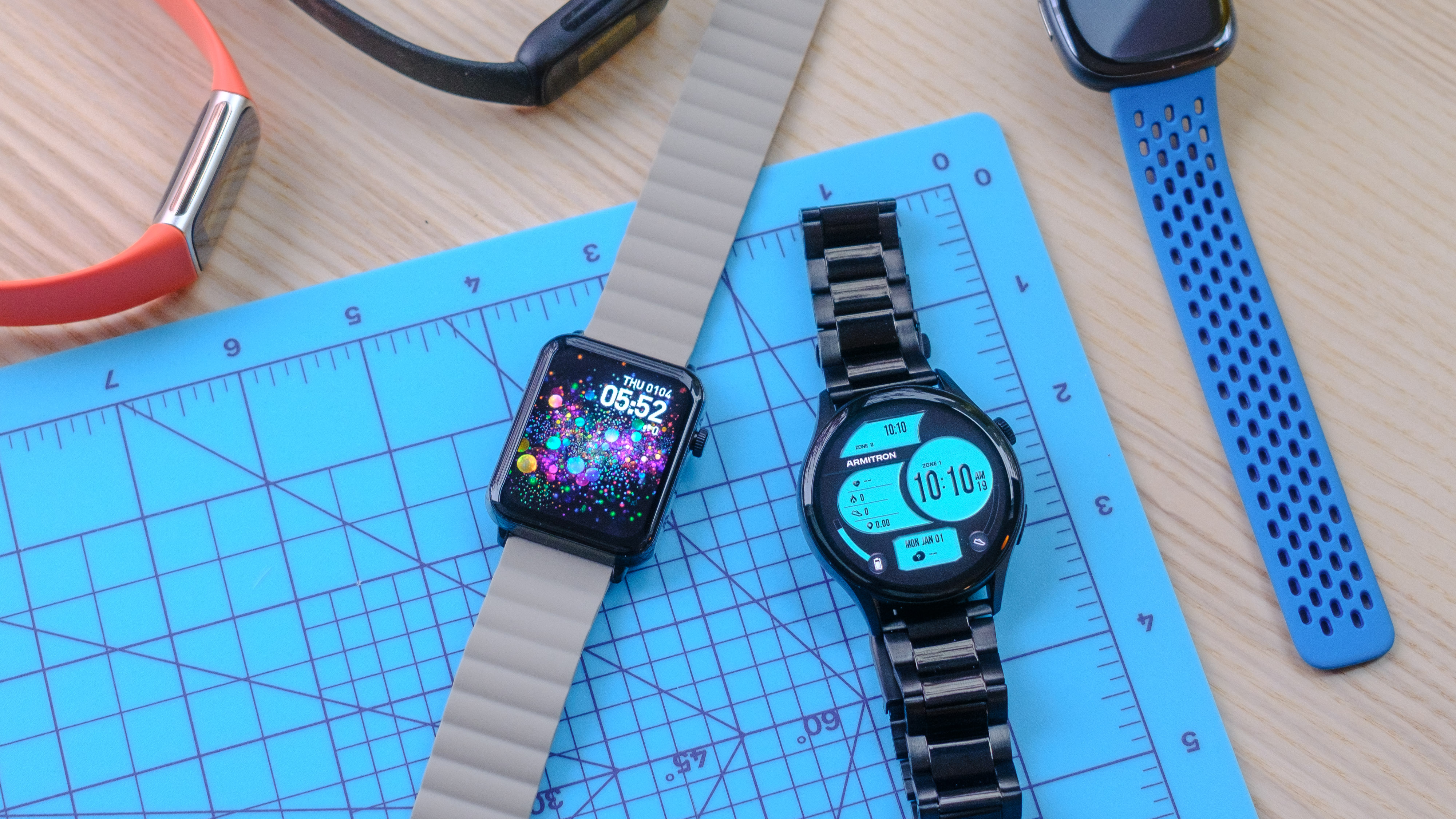 Deux modèles de montres intelligentes Armitron sur un tapis de découpe bleu avec plusieurs modèles Fitbit sur le côté de l'image