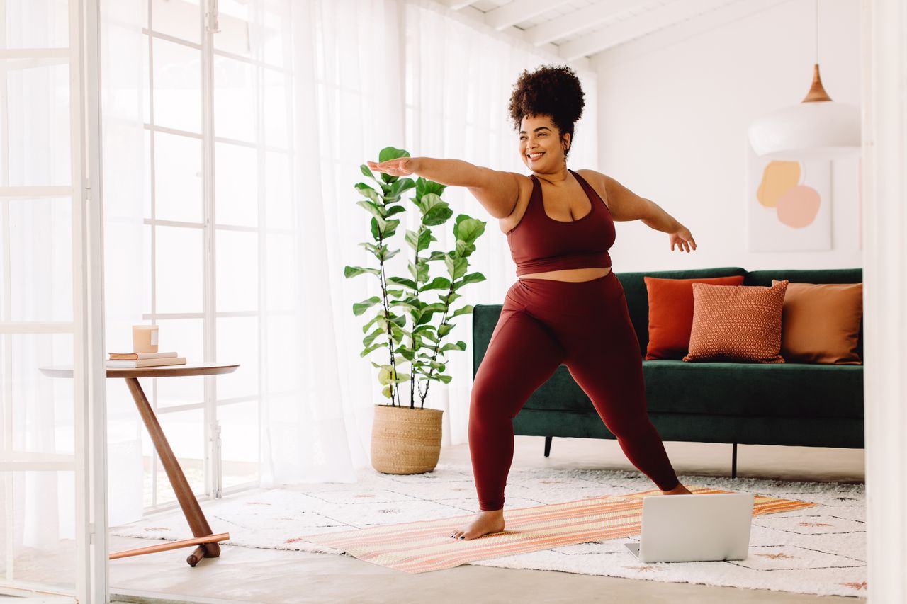 Full length of healthy woman exercising at home watching online video on laptop. Beautiful female in sports wear doing yoga.