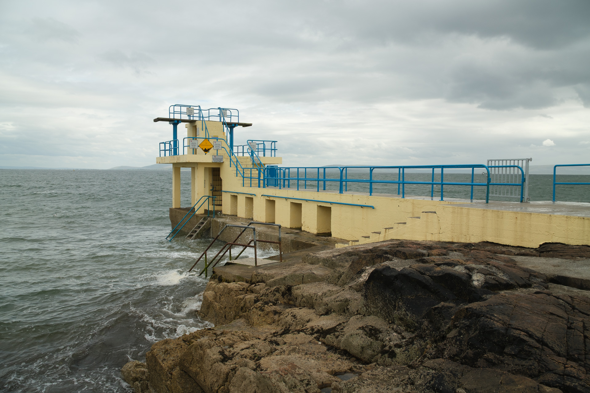 A cream diving platform with blue railings stands above a rocky shoreline and gray-green sea.