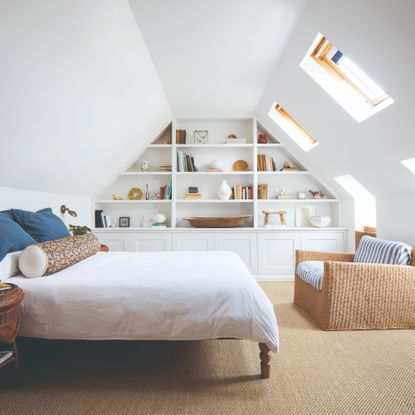 A white bedroom with slanted walls and built-in shelving, blue cushions on the bed and a rattan chair with striped blue and white cushions