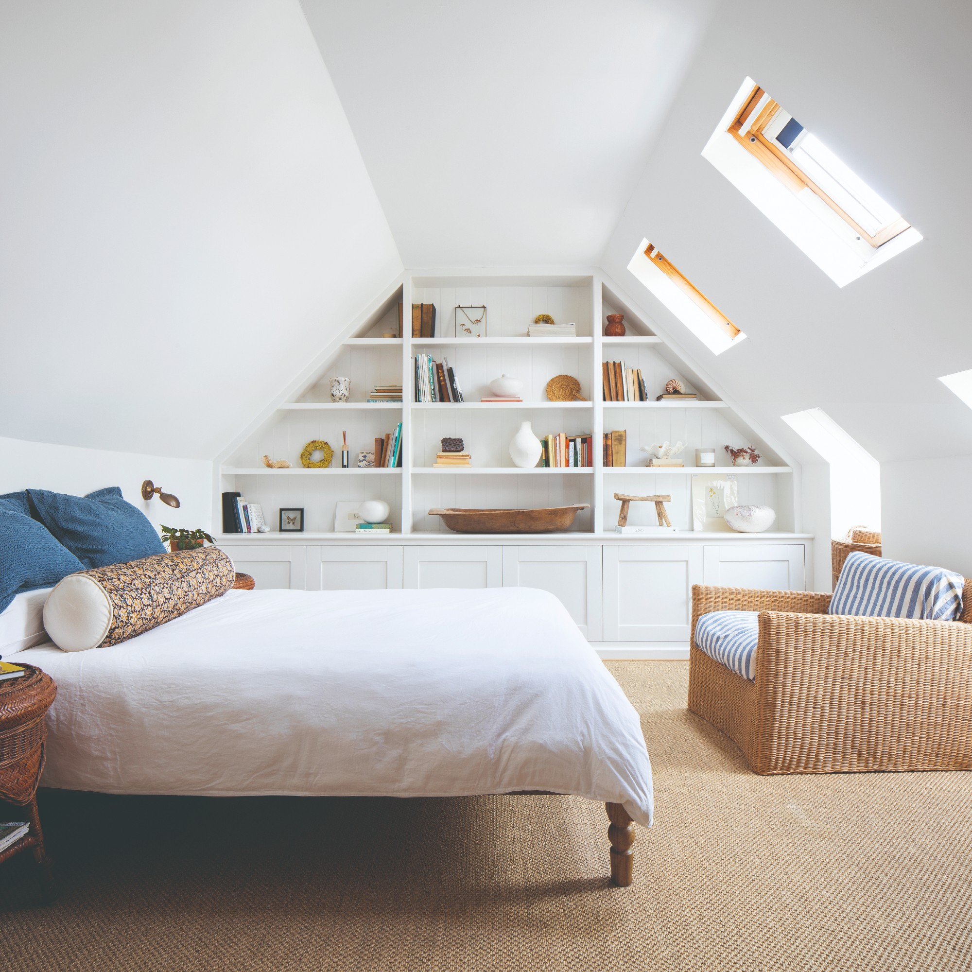 A white bedroom with slanted walls and built-in shelving, blue cushions on the bed and a rattan chair with striped blue and white cushions
