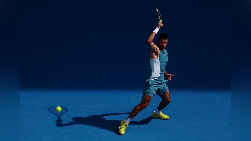 A tennis player in a turquoise outfit prepares to swing a racket on a blue court, with a ball in mid-air. Bright spotlighting creates shadows