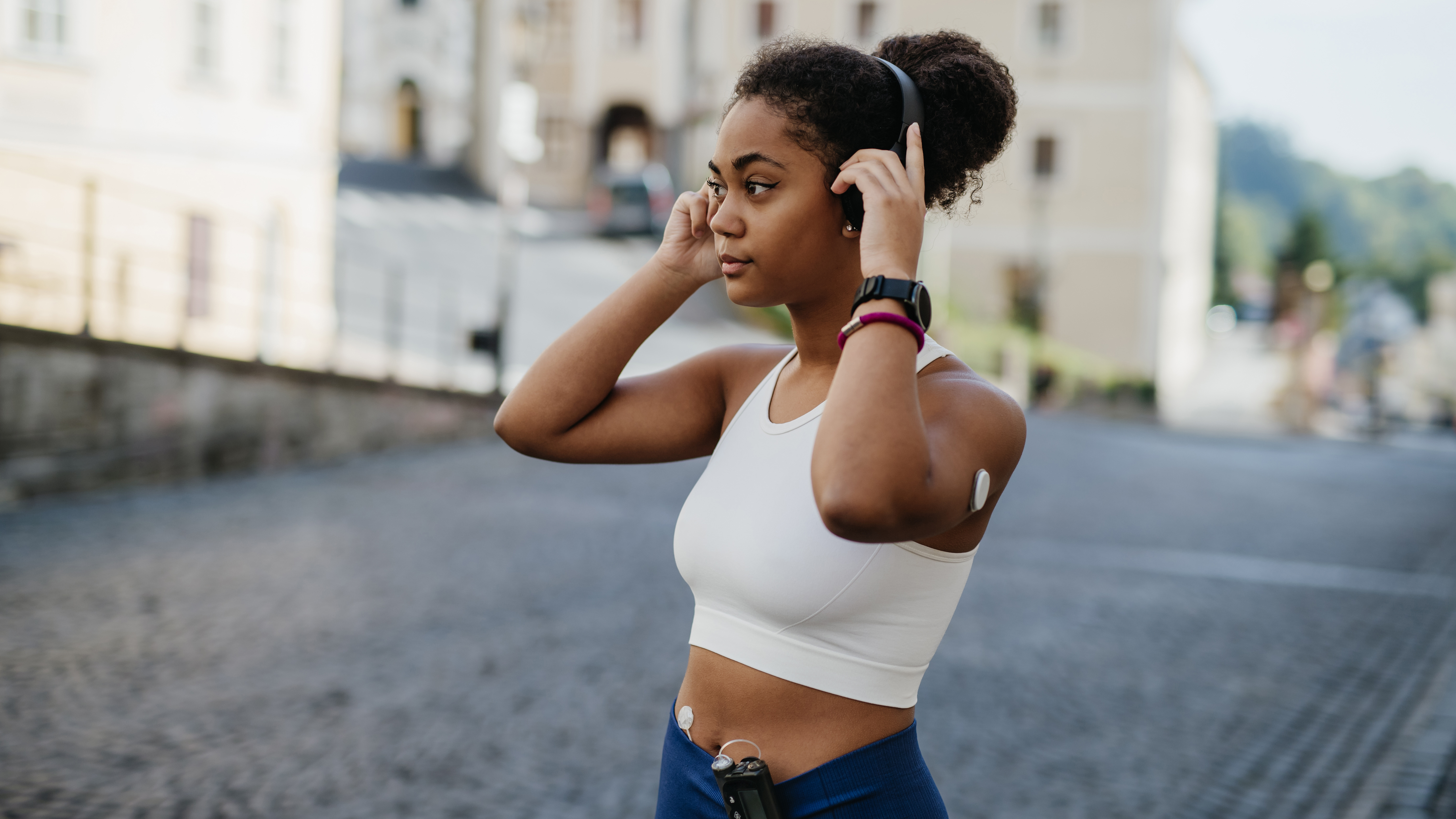 A woman in sportswear adjusts her headphones. She is wearing a glucose monitor and insulin pump.