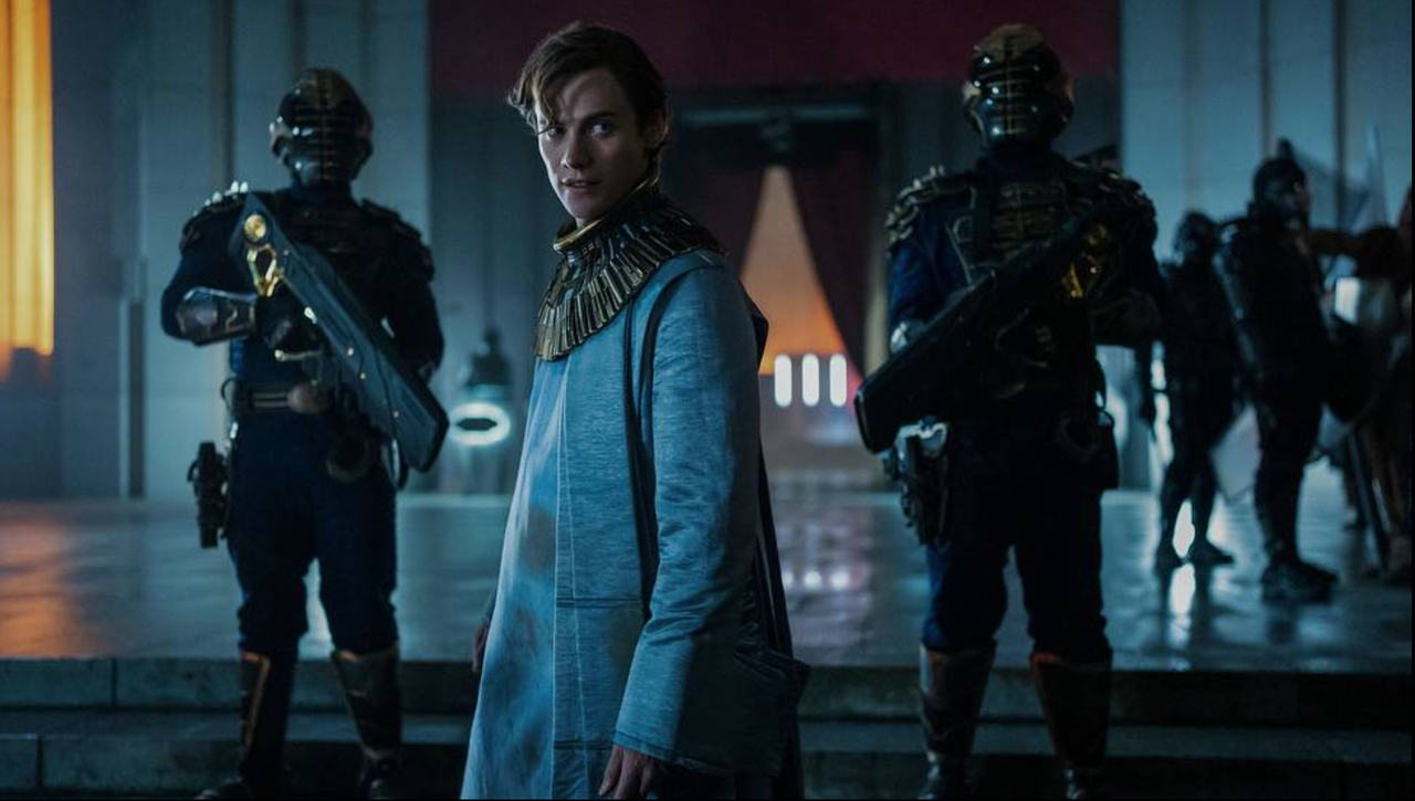 A man in a blue tunic robe stands with two armed guards
