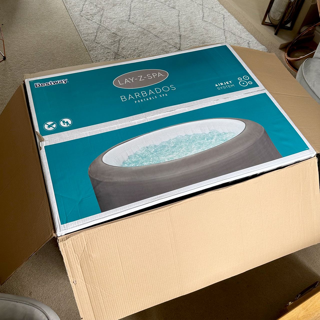 The Lay-Z-Spa Barbados inflatable hot tub in a cardboard box with a picture of the hot tub on the outside in a carpeted living room