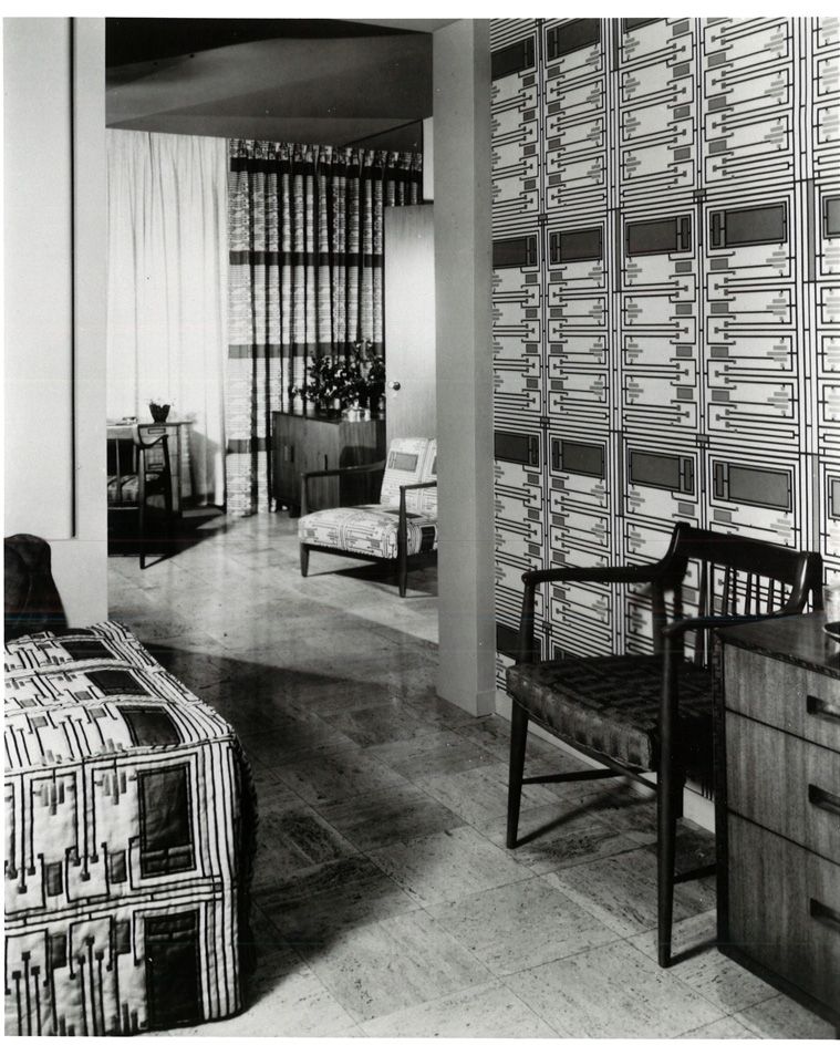 A black and white photo of a bedroom with geometric patterned fabric designs on the bed, walls and chairs.
