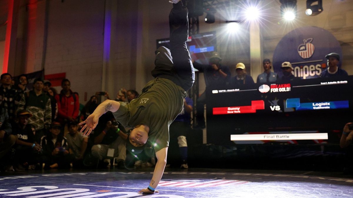 a b-girl wearing a green t shirt, black pants and a black ball cap performs a freeze on one hand in front of judges and an audience