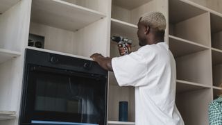 Smiling woman standing with instruction manual near man using drill on rack at home