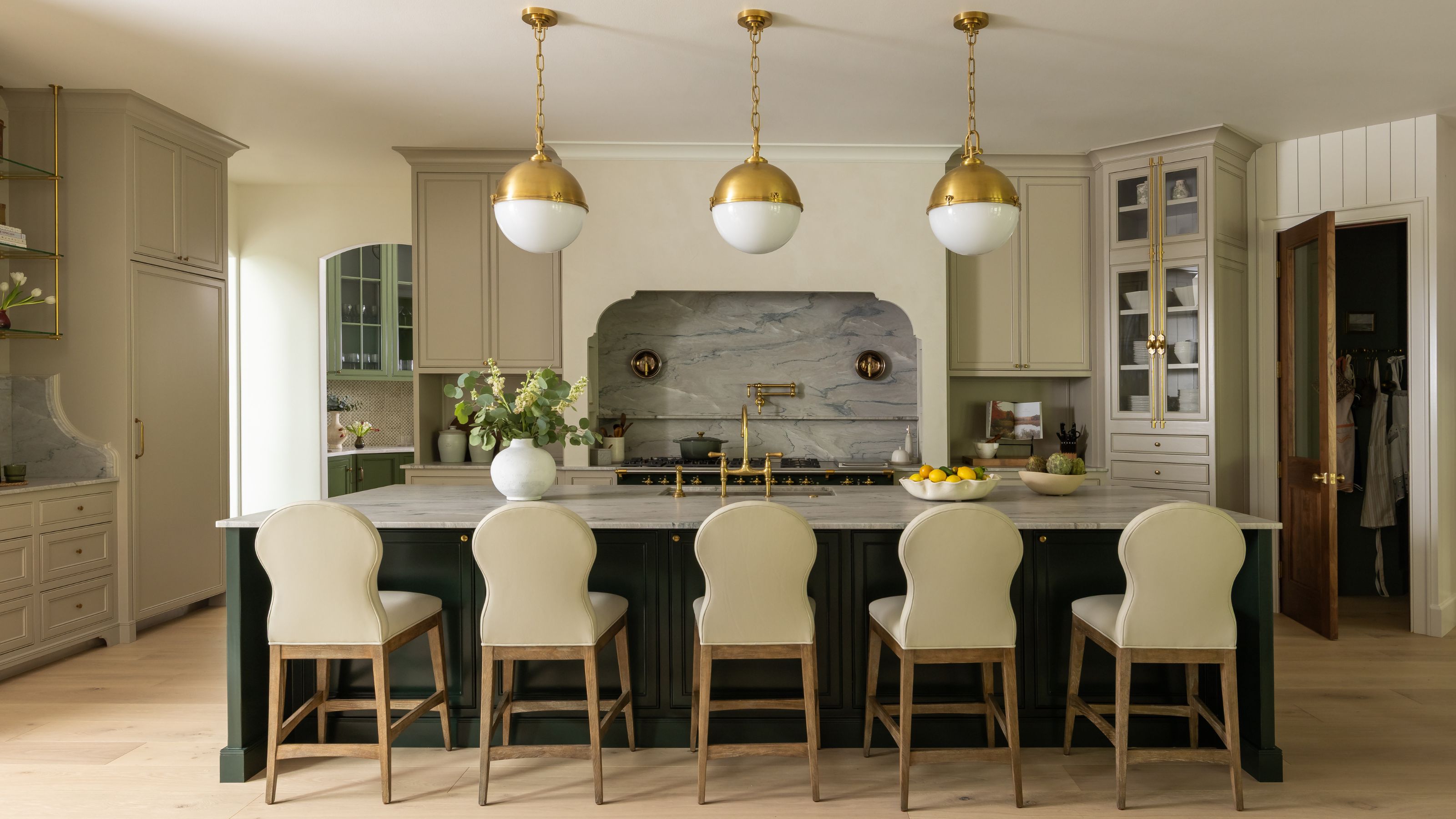 a large neutral kitchen with dark painted island and traditional bar stools, brass pendants and a curved plaster cooker hood with statement marble