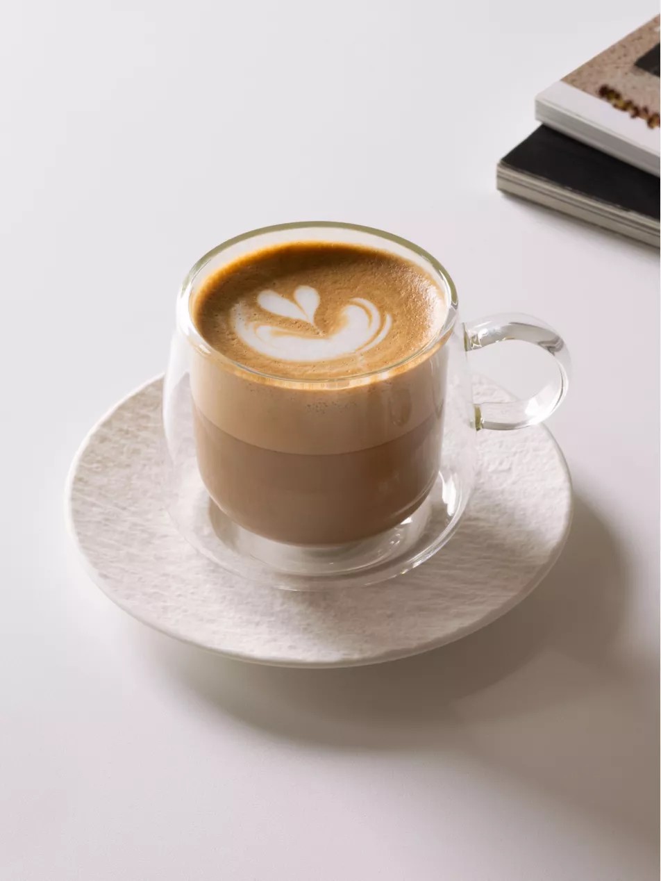 Close-up image of a glass coffee mug on a white ceramic plate that is sitting on a white countertop. The corners of two books are in view.
