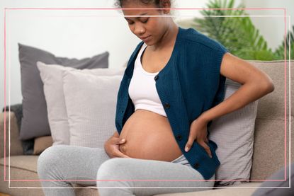 A pregnant woman sits on a sofa with one hand on her bump and the other on her lower back