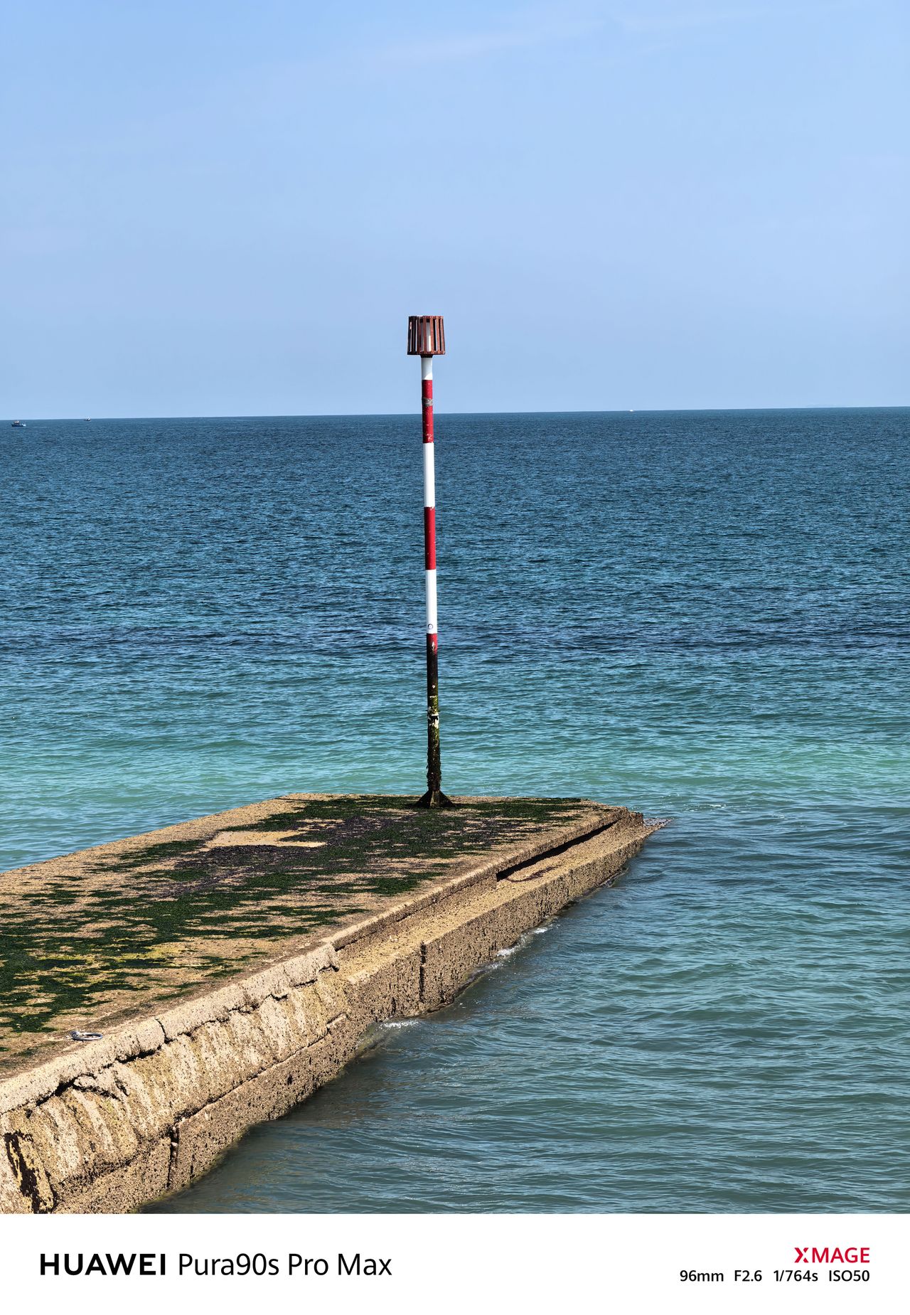 Red-and-white navigation marker at the end of a weathered concrete groyne surrounded by blue water.