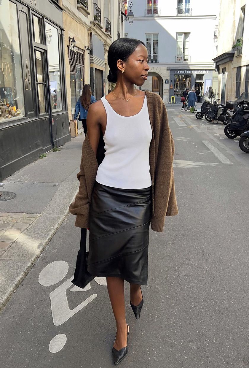 A French woman walking down the street wearing a brown cardigan styled with a white tank, black leather pencil skirt, black pumps, and a black bag.