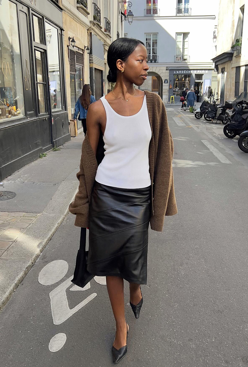 A French woman walking down the street wearing a brown cardigan styled with a white tank, black leather pencil skirt, black pumps, and a black bag.