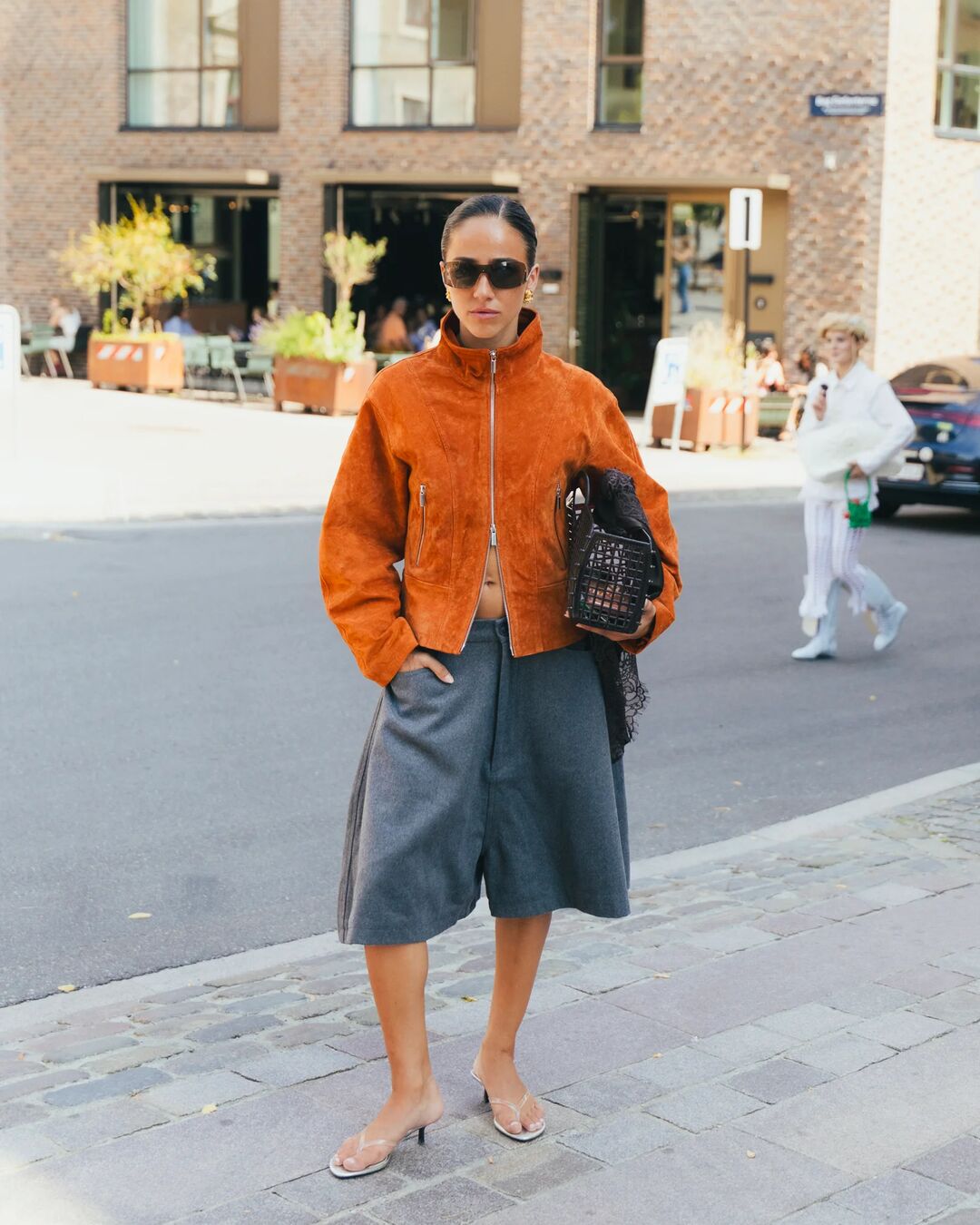 Woman wears orange funnel neck jacket and voluminous gray shorts with a clear kitten-heel.