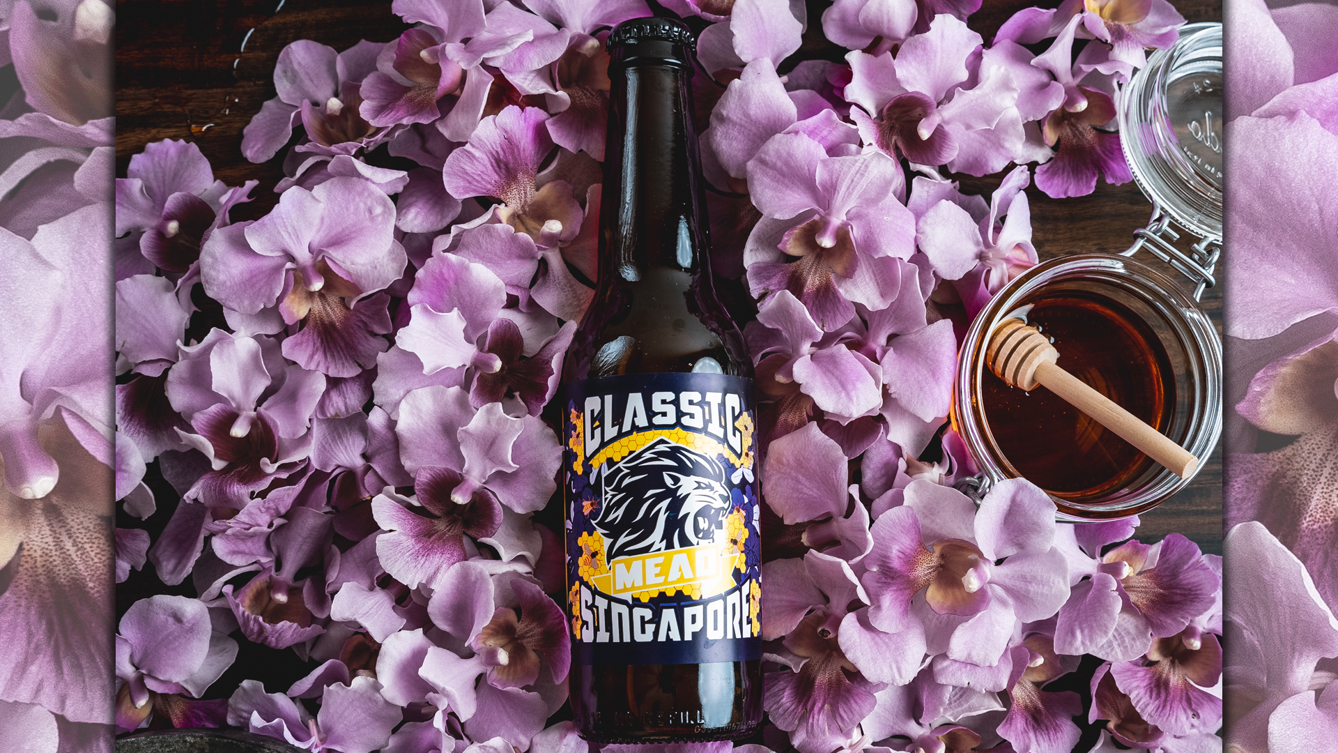A bottle of mead laid on flower petals and next to a jar of honey