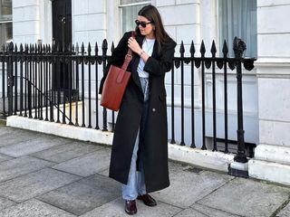 British influencer Anna Newton poses on a London sidewalk wearing a winter denim outfit with black sunglasses, a long black coat, white t-shirt, straight-leg jeans, leather tote bag, and patent burgundy loafers