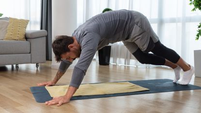 man wearing grey workout clothes performing a downward facing dog pedal stretch on an exercise mat in a living room. 