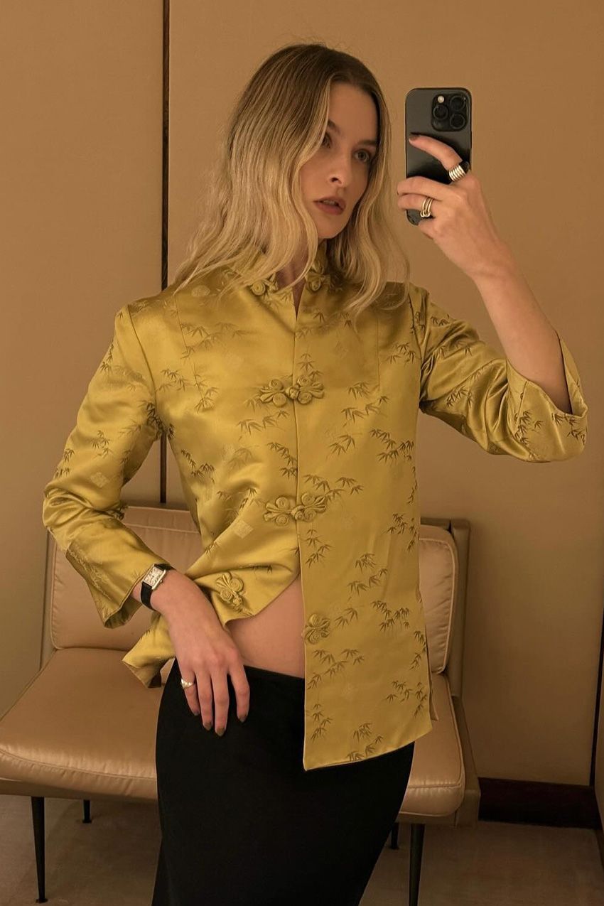 A woman taking a selfie in a mirror wearing a yellow tang jacket styled with black trousers, a black watch, and stacked rings.