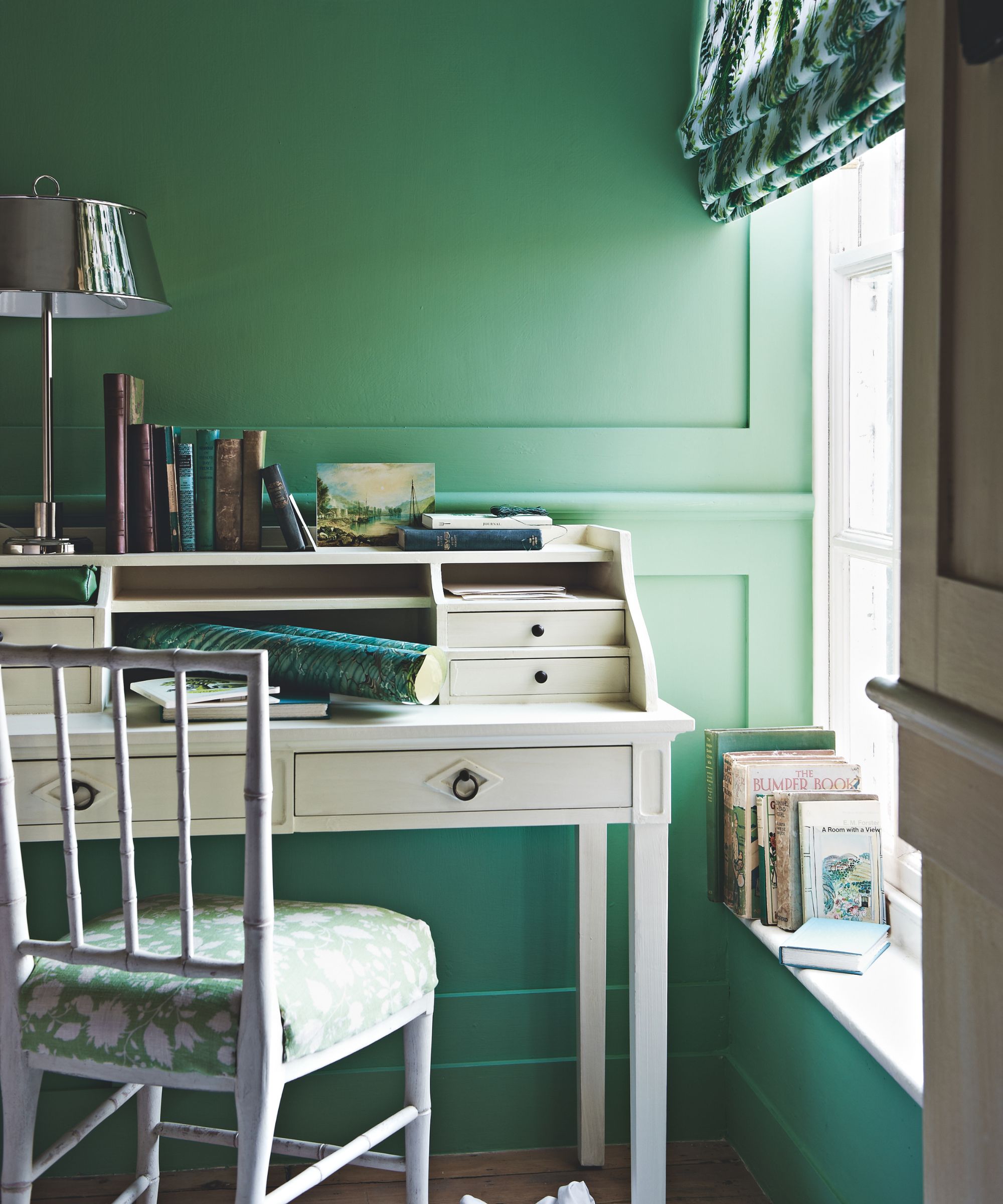White desk and chair in green painted room by a window with books on the ledge