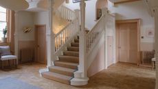 Image of a large stairwell in the middle of a large entryway. The railing is white and there is a jute stair runner. The trim on the walls is painted a dusty pink and so are the doors in the room.