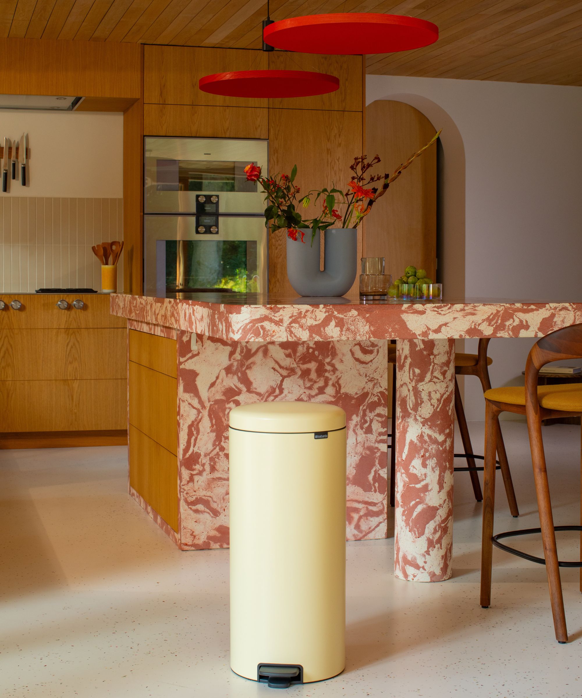 A marble and wood kitchen with a butter yellow trash can in the foreground, red marble countertops and wooden cabinets.