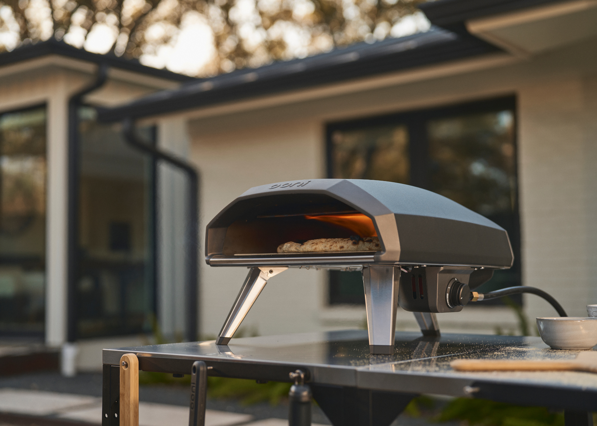 ooni koda 2 pizza oven on an outdoor surface cooking a pizza during the day with a modern home in the background