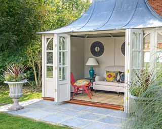 Garden shed ideas showing a smart, cream shed with leaded roof and a small gray patio in front.