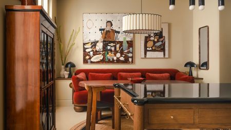 A cozy, eclectic dining area featuring a plush red velvet banquette against a beige wall. A large abstract painting of a dinner scene hangs above the table, flanked by modern black table lamps and a tiered white pendant light.