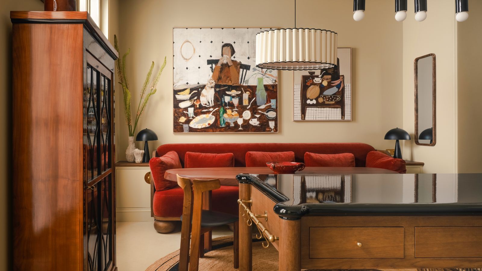A cozy, eclectic dining area featuring a plush red velvet banquette against a beige wall. A large abstract painting of a dinner scene hangs above the table, flanked by modern black table lamps and a tiered white pendant light.