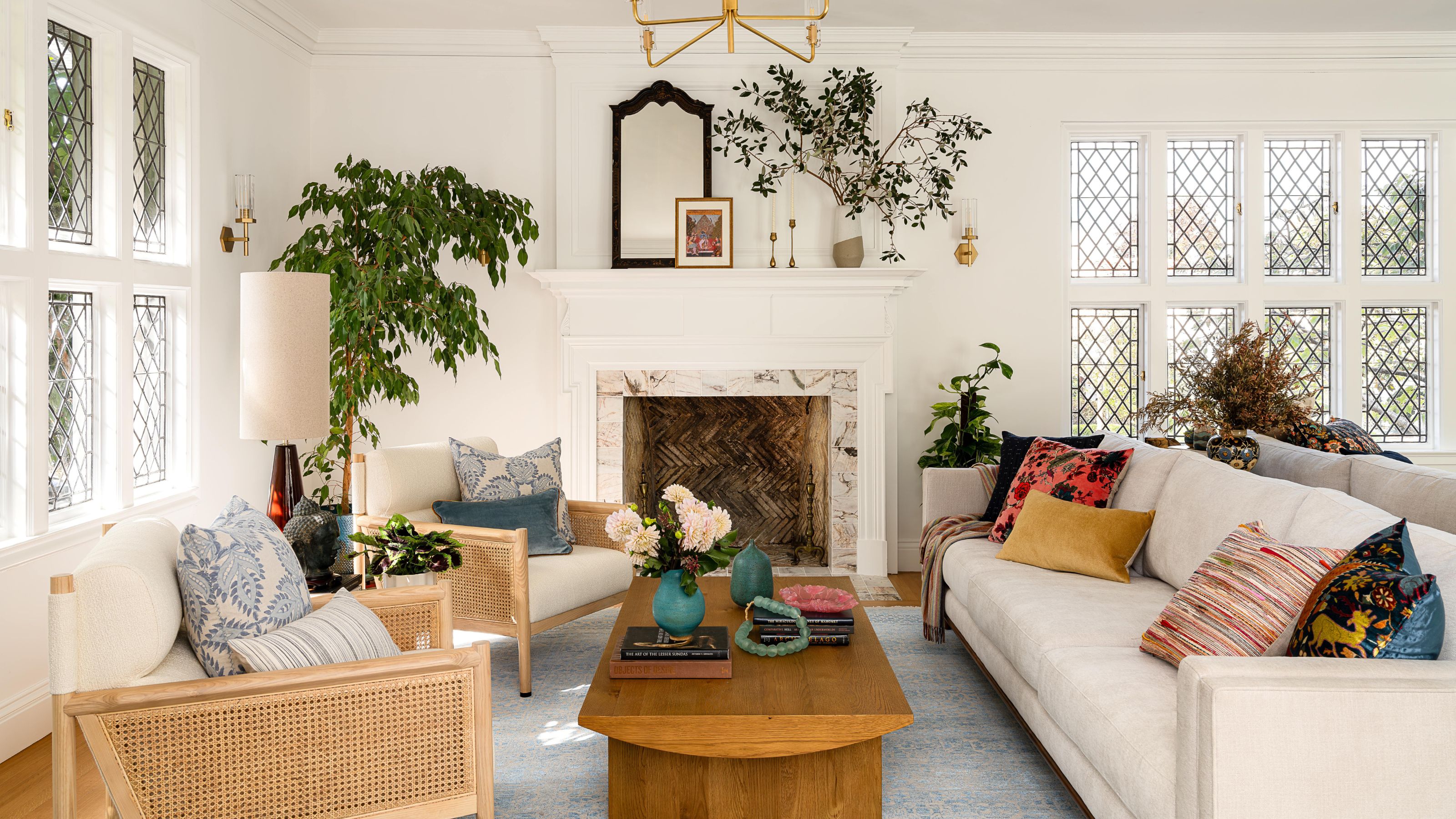 a white bright living room with original leaded tudor windows and fireplace, a neutral couch and rattan chairs with lots of house plants and a wooden coffee table