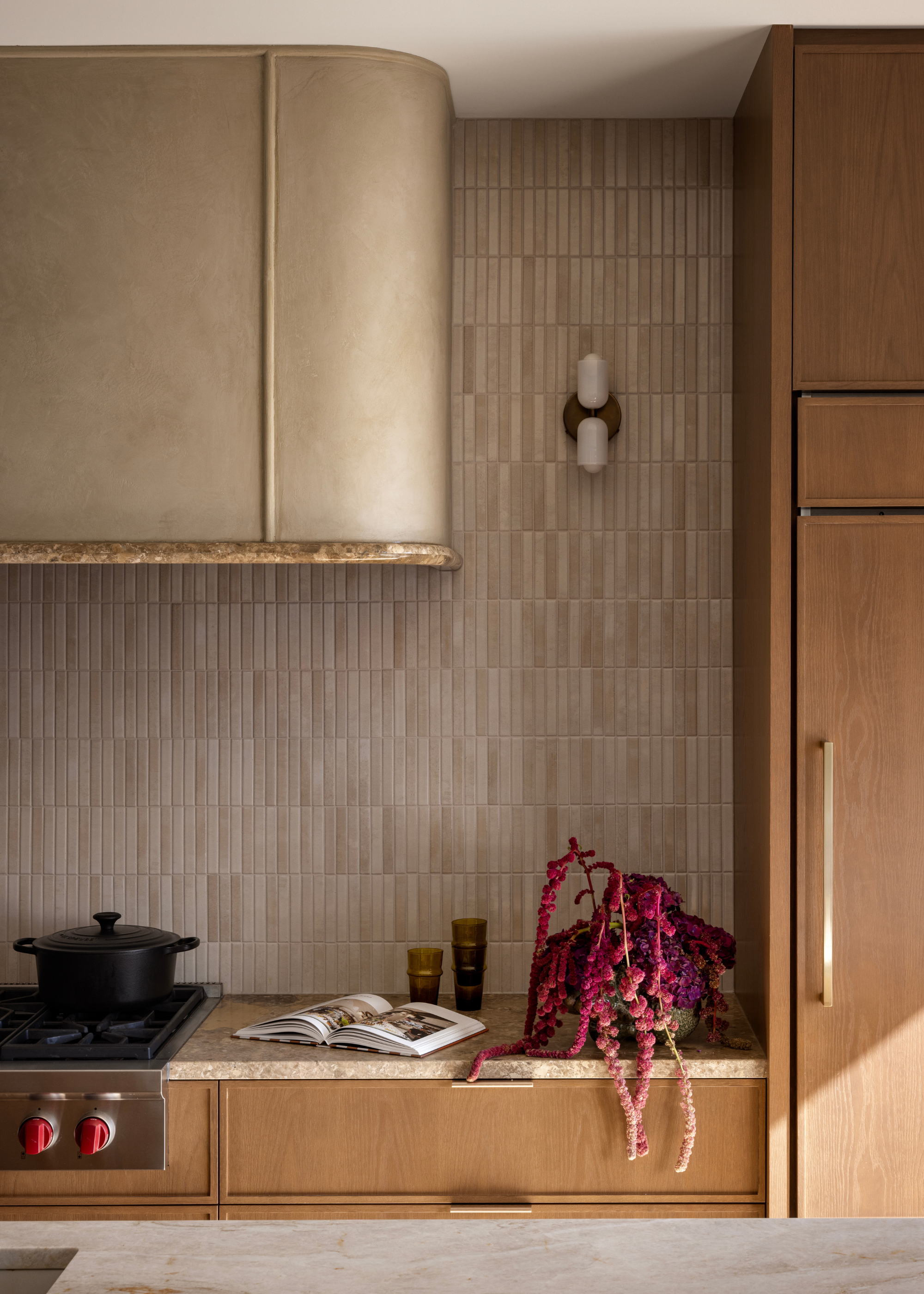 A kitchen with wood cabinets, a cream marble counter, a tiled backsplash, an extractor hood, a stove with a crock pot beside an open recipe book, a stack of glasses, and a vase of pink stems