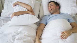 A man in a blue t-shirt snores next to his partner in bed who covers her face with a pillow