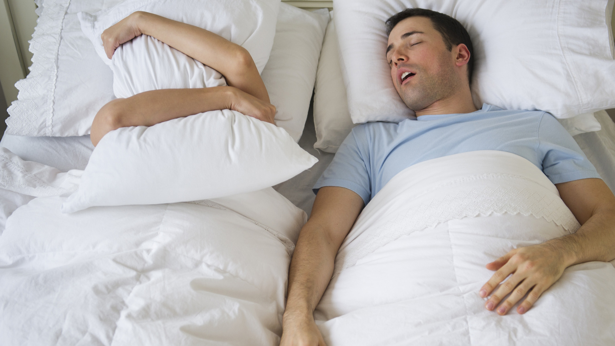 Мужчина в синей футболке храпит рядом со своей партнершей в постели, которая закрывает лицо подушкой.