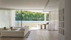 A white living room with a glass door to the garden and outdoor dining space. White tiles hung on the wall, a large couch on a large rug, a coffee table and a side table with a vase