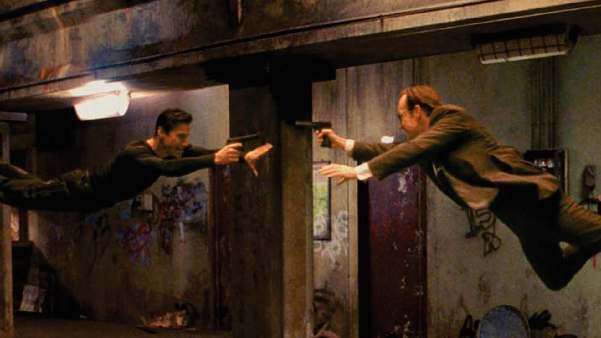 A still from The Matrix movie. Set in a dirty and graffitied underground train station, there are two men flying towards each other, each man pointing a gun at the other. The man on the left is Neo, dressed in black jeans and a black long-sleeved shirt. On the right is Agent Smith, dressed in a suit.