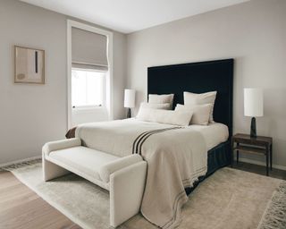 Neutral contemporary bedroom, gray painted walls, dark wood flooring with rug, large bed with black upholstered headboard, cream bedding and cushions, boucle upholstered daybed at foot of bed
