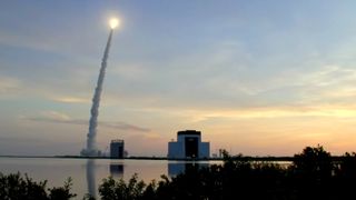 a rocket launches into the early morning sky in the distance on the left leaving a cloudy trail behind it, some buildings are visible in the distance.