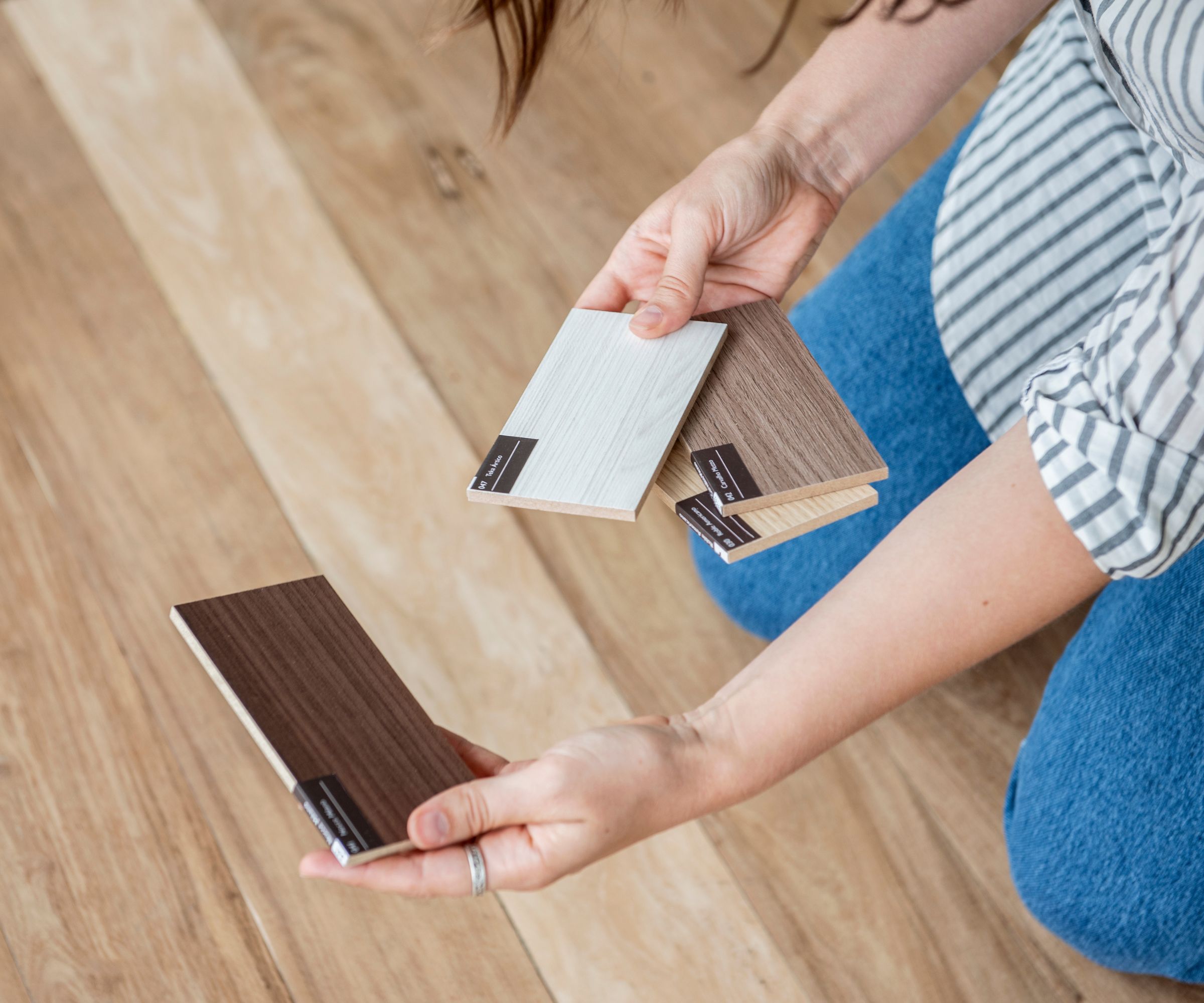 Hands comparing wood flooring samples over natural timber planks, showcasing light, mid, and dark tones to help choose the perfect floor finish.