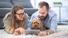 A Millennial couple looks at a tablet on their living room floor with their dog.