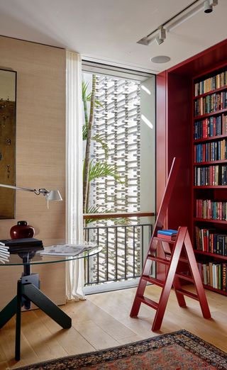 A red ladder placed by a red filled floor to ceilingbook shelf. Floor to ceiling glas window. Round glass table