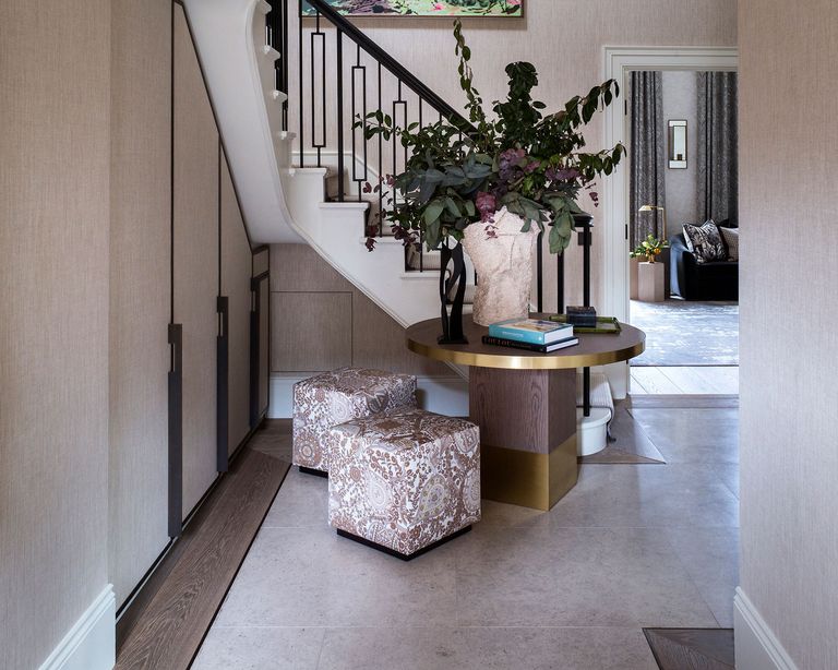 Narrow hallway ideas shown in a beige scheme with round gilt-edged table and square footstools.
