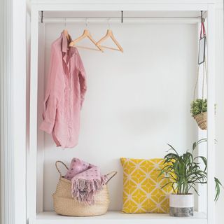 An open wooden wardrobe with a shirt and other hangers hanging from it, there's a wicker basket and a yellow cushion underneath