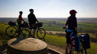 Three cyclists are silhouetted and overlooking a green landscape from a lookout point
