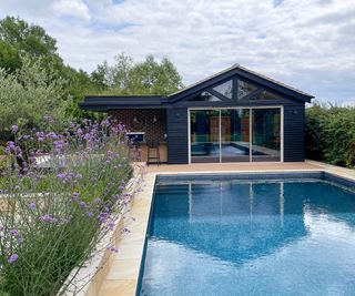 A pool area surrounded with minima stone paving with substantial planting beds containing tall perennial plants which provide a screen. The pool house in the background is in dark timber with large glazing